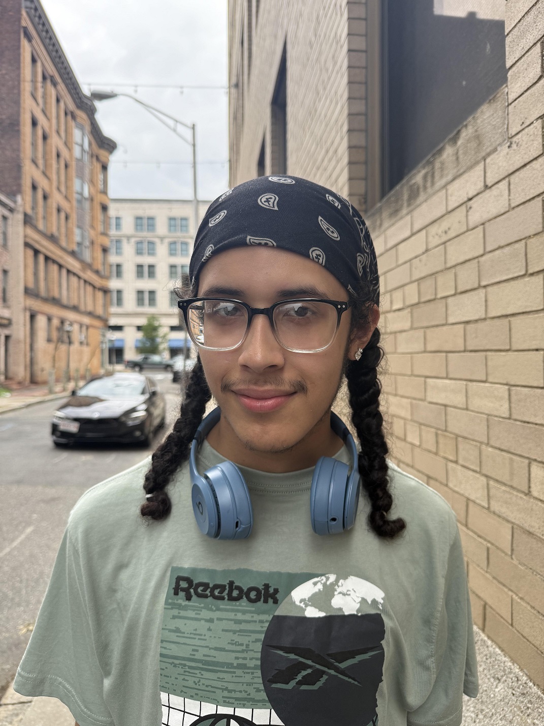 A young man with glasses, bandana, braids, headphones smiling outside of a brick building