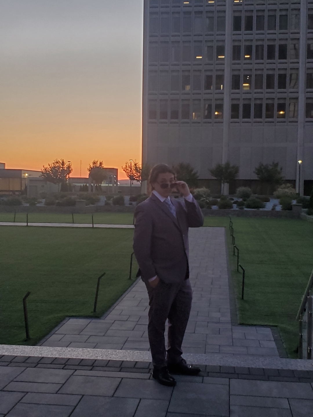 A young man in a suit and sunglasses posing in front of a sunset and a large building