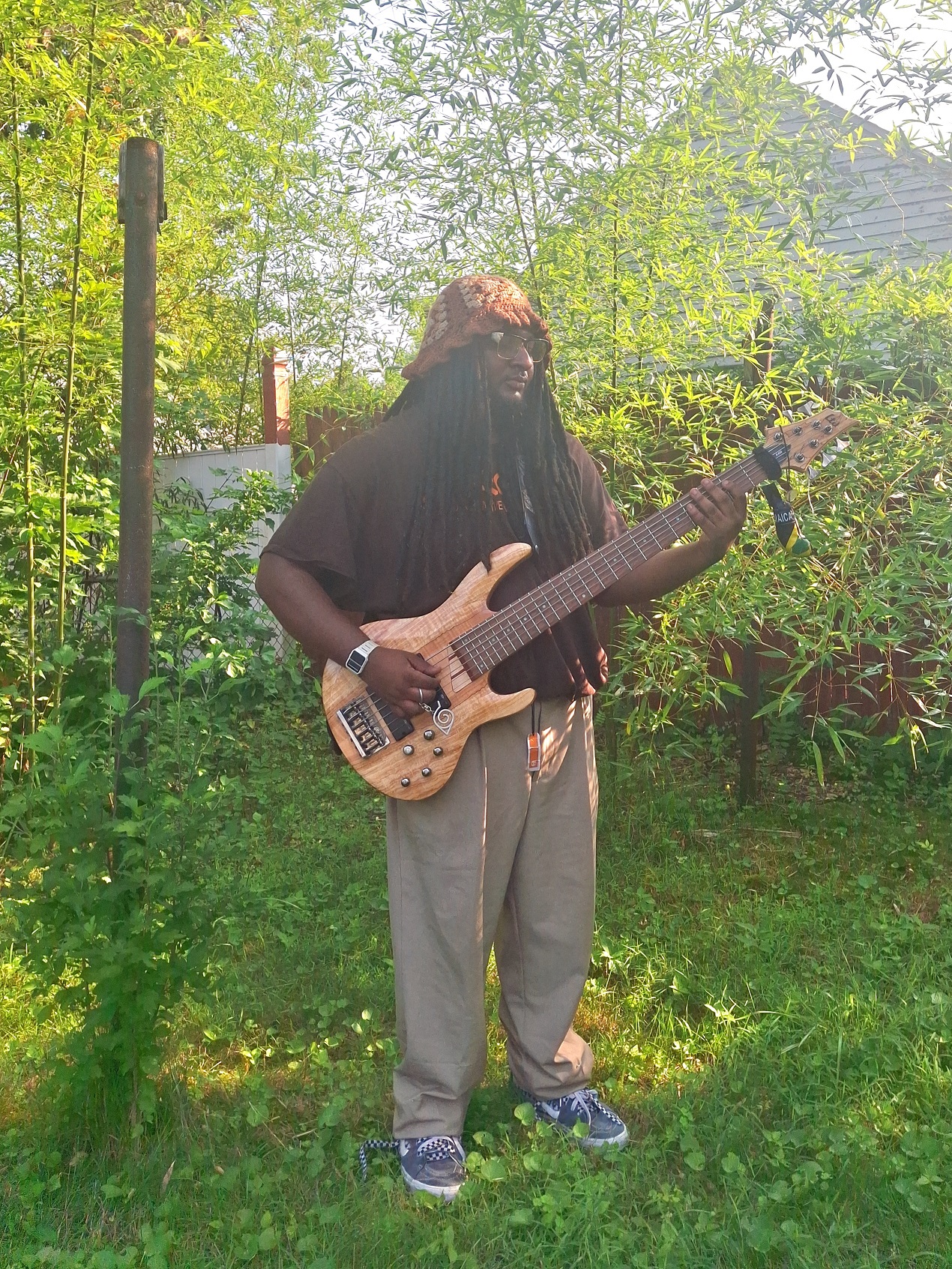 A young man surrounded by trees playing bass, with long dreads, wearing a bucket hat