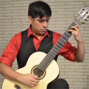 John Cesar playing guitar in front of white brick, wearing a red button up with black vest, looking down