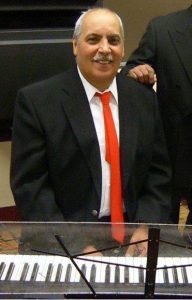 Joe Velez sitting behind a keyboard wearing a black suit and red tie, smiling