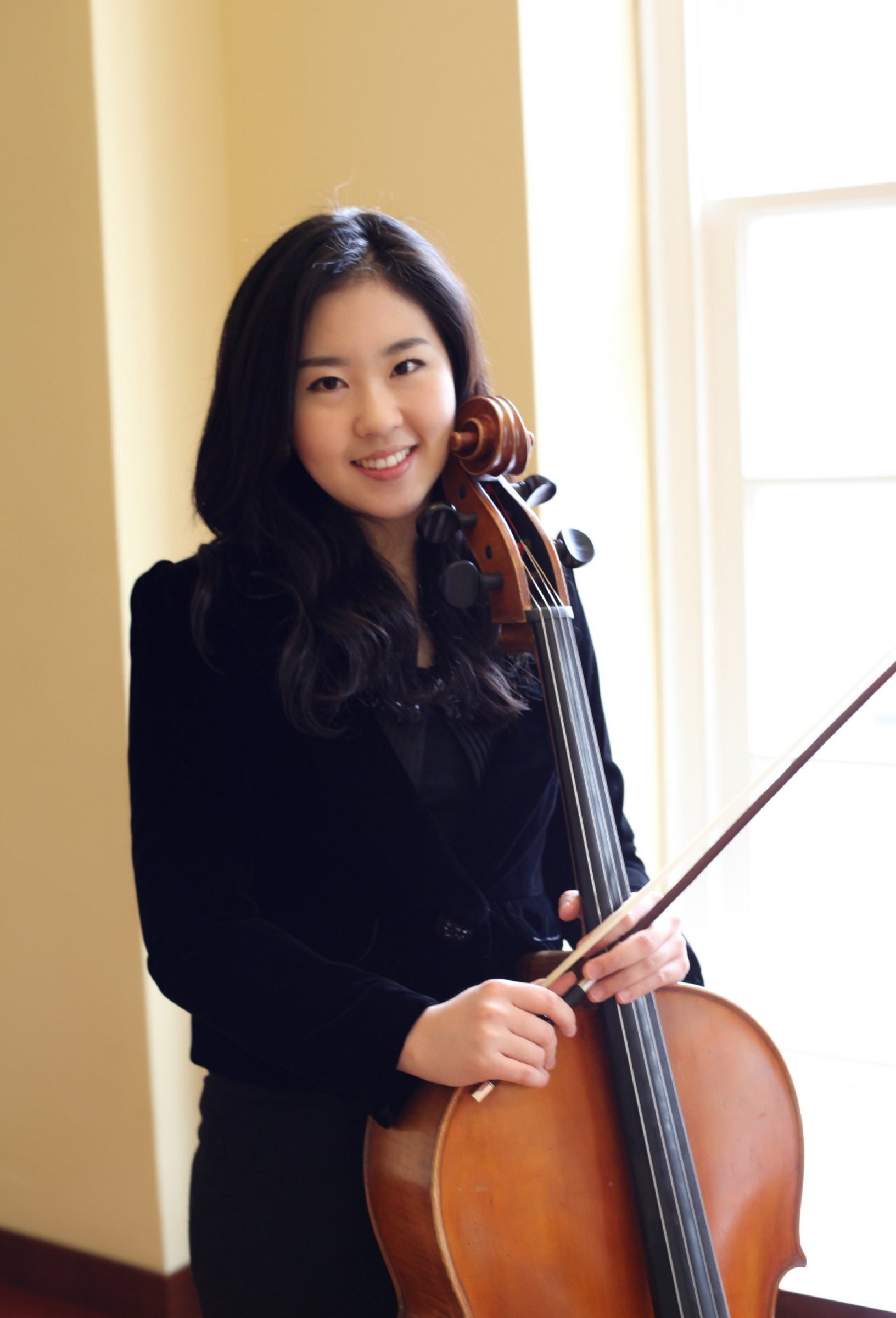 Yoonhee Ko posing and smiling with her cello in front of sunny window