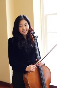 Yoonhee Ko posing and smiling with her cello in front of sunny window