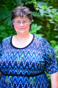 Headshot of Joan Marcil, smiling outside surrounded by green leaves, showing from the hips and above in a blue blouse