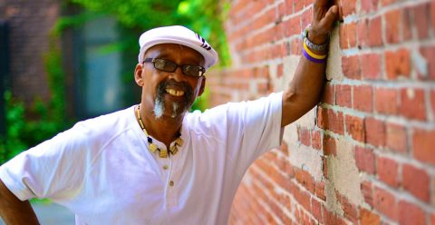 Headshot of Billy Arnold leaning on a brick building outside, wearing a white shirt and hat smiling