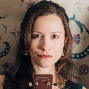 Headshot of Liz Simmons, against flower-patterned wall, showing shoulders and up with top of a guitar on the bottom of picture