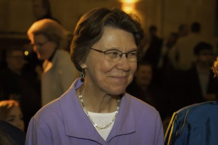 Headshot of Mary Ellen Miller smiling and looking to the side in a group of people, wearing a purple blouse, blurred background