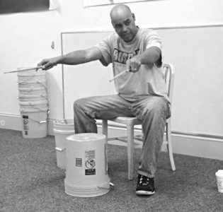 Rick Marshall teaching a bucket drum lesson with his arms stretched out in front holding drum sticks