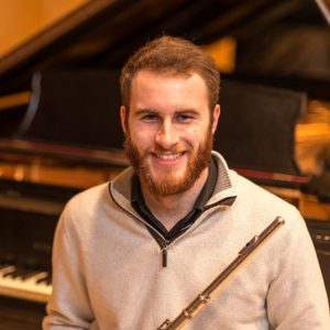 Headshot of Zach Robarge sitting at a piano with his flute smiling