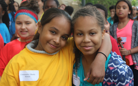 Two students posing, one with their arm around the other smiling, other students blurred in the background