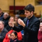 Chris Moehringer conducting YWES in a suit at their winter concert, blurred audience members in the background