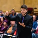 Chris Moehringer conducting YWES and smiling in a suit at their winter concert, blurred audience members in the background