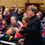 Assistant conductor of YWES leading them in their winter concert, blurred audience members in the background