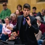 Chris Moehringer conducting YWES in a suit at their winter concert, blurred audience members in the background