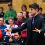 Chris Moehringer conducting YWES and making an intense face in a suit at their winter concert, blurred audience members in the background
