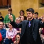 Chris Moehringer conducting YWES and smiling in a suit at their winter concert, blurred audience members in the background