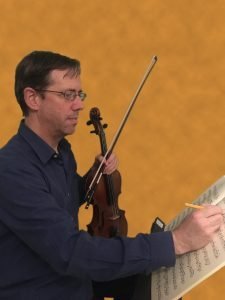 greg holding violin and bow with sheet music in front of him, writing on sheet music with a pencil