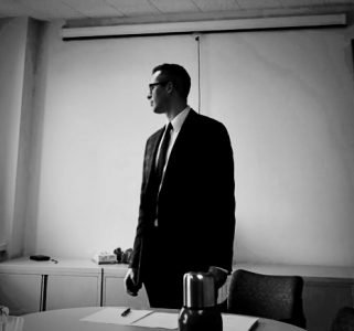black and white photo of Matthew Holmes wearing a suit and tie, standing and looking to his right in front of a classroom