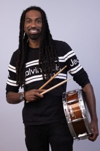 A man with long black twisted dreadlocks and a calvin klein shirt standing, smiling, holding 2 drumsticks in one hand and a drum at his side in the other hand