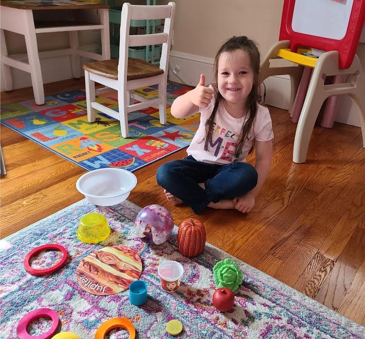 Prelude student sitting with toys, cross-legged, giving a thumbs-up and smiling to the camera