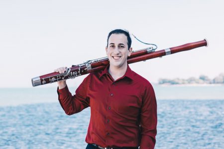 Vinny holding his bassoon over his shoulder, standing in front of a body of water
