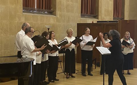 Group of adults in concert hall singing and holding folders with conductor directing them