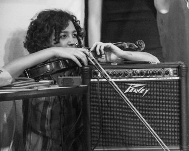 a person with curly hair holding a violin and bow, standing behind a speaker and resting their arms on top of the speaker