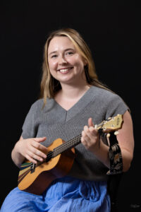 a woman with long light brown hair smiling strumming a ukulele