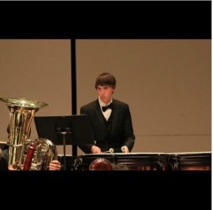 a young man with light skin and brown hair plays orchestral percussion while looking at a music stand; a sousaphone is visible to his left