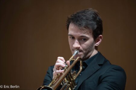 a young man with light skin and brown hair playing a trumpet wearing a black collared jacket