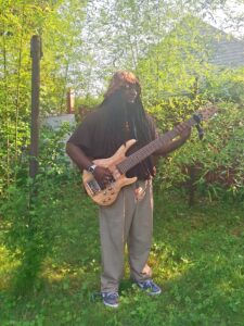A young man surrounded by trees playing bass, with long dreads, wearing a bucket hat